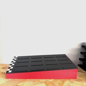May include: A red and black display stand with a checkered pattern on one corner. The top surface is black with rectangular grooves. The stand is on a wooden surface.