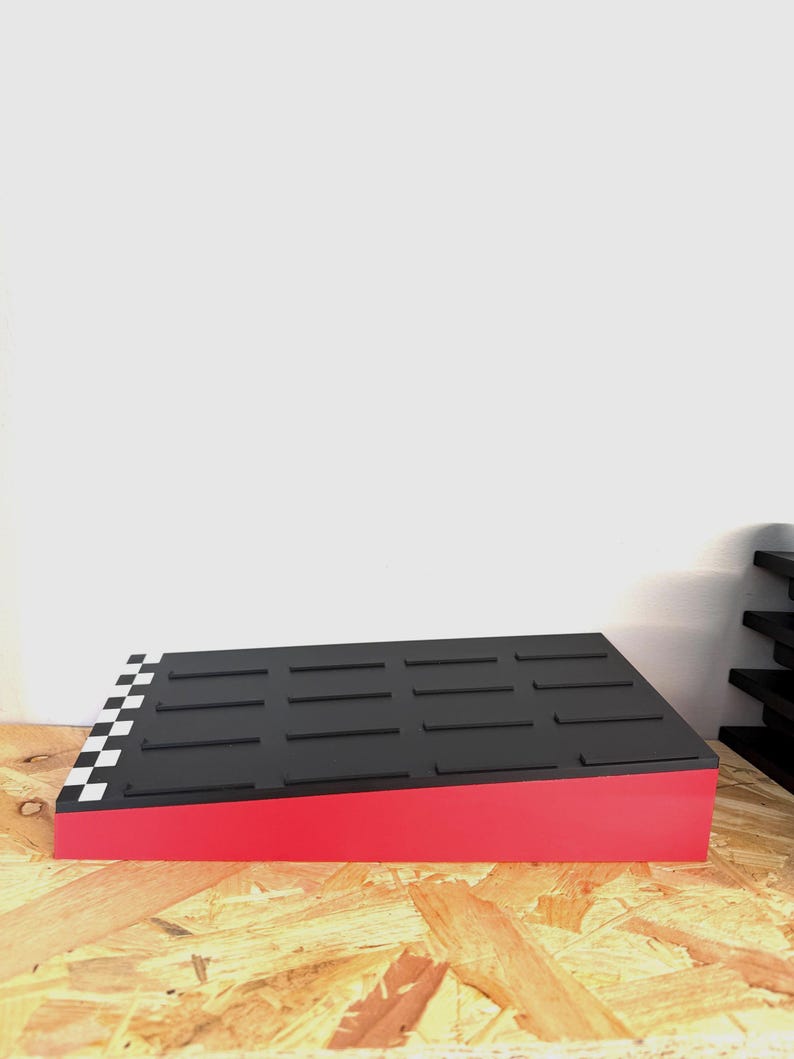 May include: A red and black display stand with a checkered pattern on one corner. The top surface is black with rectangular grooves. The stand is on a wooden surface.