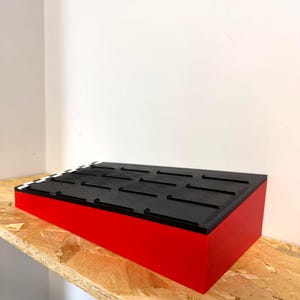 May include: A red and black angled display stand. The stand has a black top with evenly spaced, raised black lines. The sides are a bright red. The stand is placed on a wooden shelf against a white wall.