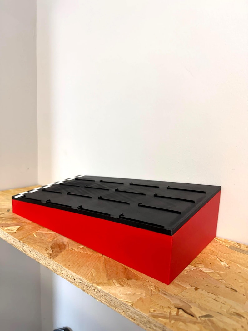 May include: A red and black angled display stand. The stand has a black top with evenly spaced, raised black lines. The sides are a bright red. The stand is placed on a wooden shelf against a white wall.