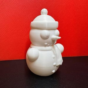 White 3D-printed snowman LED tealight holder shown against a bold red background, highlighting its intricate winter-themed details like the scarf, buttons, and hat.