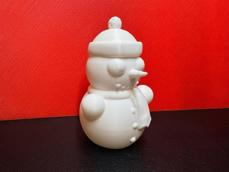 White 3D-printed snowman LED tealight holder shown against a bold red background, highlighting its intricate winter-themed details like the scarf, buttons, and hat.
