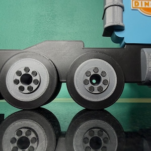 May include: A close-up of a toy truck's wheels and chassis. The wheels are black with gray centers and detailed bolt patterns. The truck's body is blue with the Dinoco logo visible. The image is set against a green background.