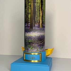 May include: A cylindrical container featuring a forest scene with bluebell flowers, mounted on a light blue square base. The base includes a gold-colored plaque with the name Cain engraved in black and two gold-colored wings extending from the sides.