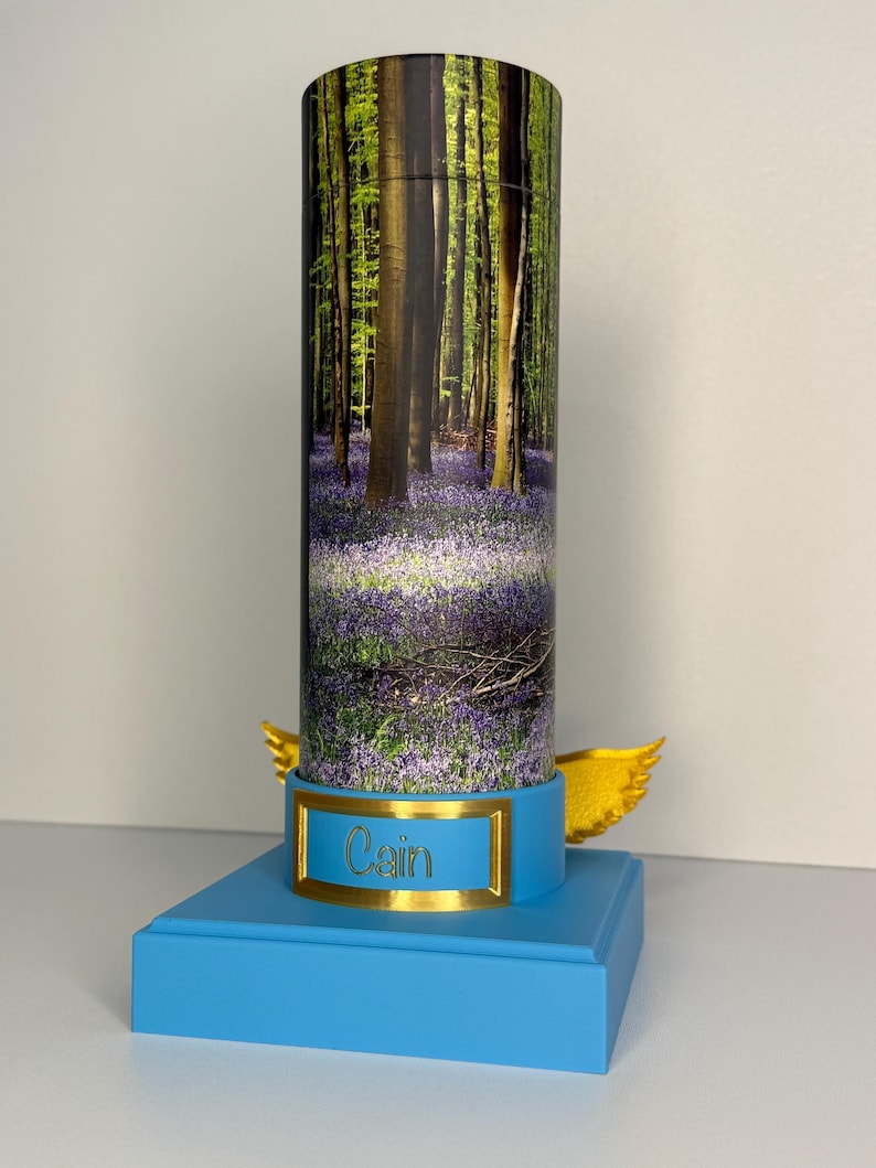 May include: A cylindrical container featuring a forest scene with bluebell flowers, mounted on a light blue square base. The base includes a gold-colored plaque with the name Cain engraved in black and two gold-colored wings extending from the sides.