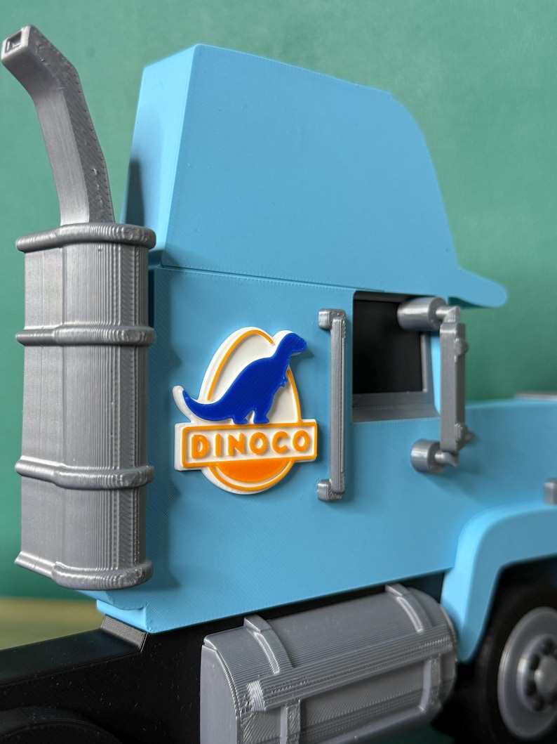 May include: A light blue toy truck with a silver exhaust pipe and fuel tank. The truck features a Dinoco logo with a blue dinosaur on a white and orange background. The truck is a toy.