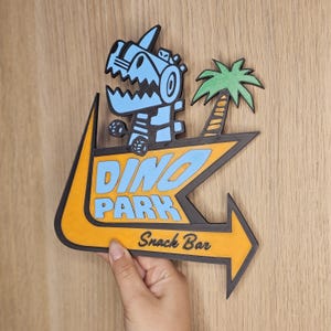 May include: A wooden Dino Park sign featuring a cartoon dinosaur and palm tree. The sign is yellow and black with blue text that reads "DINO PARK Snack Bar". The arrow points to the right.