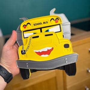 K&ouml;nnte beinhalten: Gelbe, schulbusf&ouml;rmige Dekoration mit einem l&auml;chelnden Gesicht, schwarzen Reifen und dem Schriftzug "SCHOOL BUS" oben. Der Bus hat eine silberne Sto&szlig;stange und eine Kettenverzierung. Der Hintergrund ist unscharf.