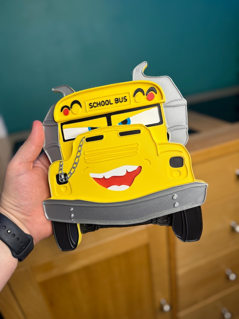 K&ouml;nnte beinhalten: Gelbe, schulbusf&ouml;rmige Dekoration mit einem l&auml;chelnden Gesicht, schwarzen Reifen und dem Schriftzug "SCHOOL BUS" oben. Der Bus hat eine silberne Sto&szlig;stange und eine Kettenverzierung. Der Hintergrund ist unscharf.