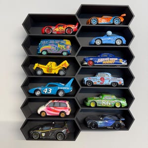 May include: A black honeycomb-shaped wall shelf containing twelve die-cast toy cars. The collection includes various colorful vehicles, such as a red number 95 race car, a blue number 43 Dinoco car, a yellow truck, and a blue Fabulous Hudson Hornet.