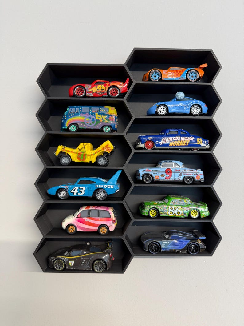 May include: A black honeycomb-shaped wall shelf containing twelve die-cast toy cars. The collection includes various colorful vehicles, such as a red number 95 race car, a blue number 43 Dinoco car, a yellow truck, and a blue Fabulous Hudson Hornet.