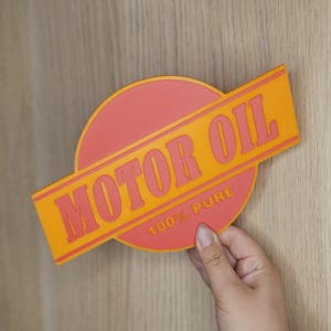 Puede incluir: Un letrero bicolor naranja y rojo con las palabras "MOTOR OIL" en letras grandes y en negrita. El letrero tambi&eacute;n incluye el texto "100% PURE" en letras m&aacute;s peque&ntilde;as. El letrero est&aacute; sostenido por una mano.