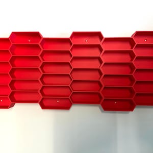 May include: A bright red wall-mounted storage organizer featuring a repeating honeycomb pattern with multiple hexagonal compartments. The structure is composed of interconnected cells arranged in a grid, providing numerous open slots for storage.