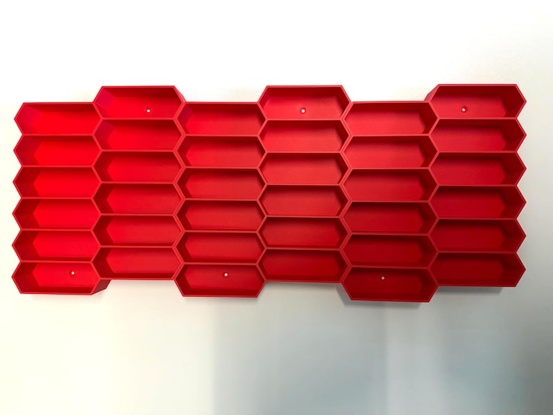 May include: A bright red wall-mounted storage organizer featuring a repeating honeycomb pattern with multiple hexagonal compartments. The structure is composed of interconnected cells arranged in a grid, providing numerous open slots for storage.