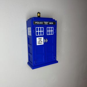 May include: A blue Police Box replica with white window frames and the text "POLICE PUBLIC CALL BOX" on top. The door has the text "FREE FOR USE OF PUBLIC". A yellow light sits on top of the box.