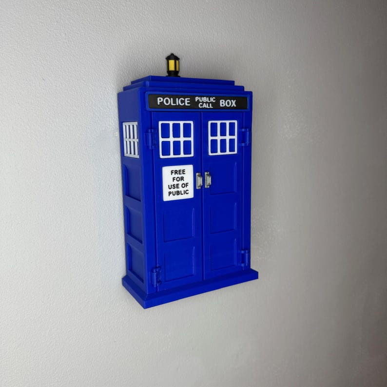 May include: A blue Police Box replica with white window frames and the text "POLICE PUBLIC CALL BOX" on top. The door has the text "FREE FOR USE OF PUBLIC". A yellow light sits on top of the box.