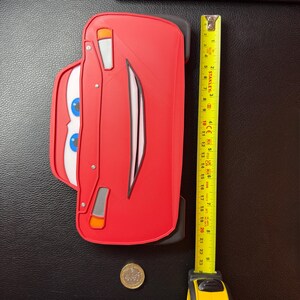 K&ouml;nnte beinhalten: Ein rotes Lightning McQueen-Auto-f&ouml;rmiges Objekt mit wei&szlig;en und blauen Augen, etwa 25 cm lang. Ein gelbes Ma&szlig;band liegt neben dem Auto. Eine M&uuml;nze befindet sich am unteren Rand des Bildes.