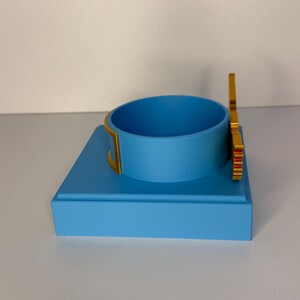 May include: A light blue square display base featuring a matching light blue circular ring holder in the center. The circular holder is accented with gold-colored trim on the sides and a vertical gold-colored decorative element attached to the back.