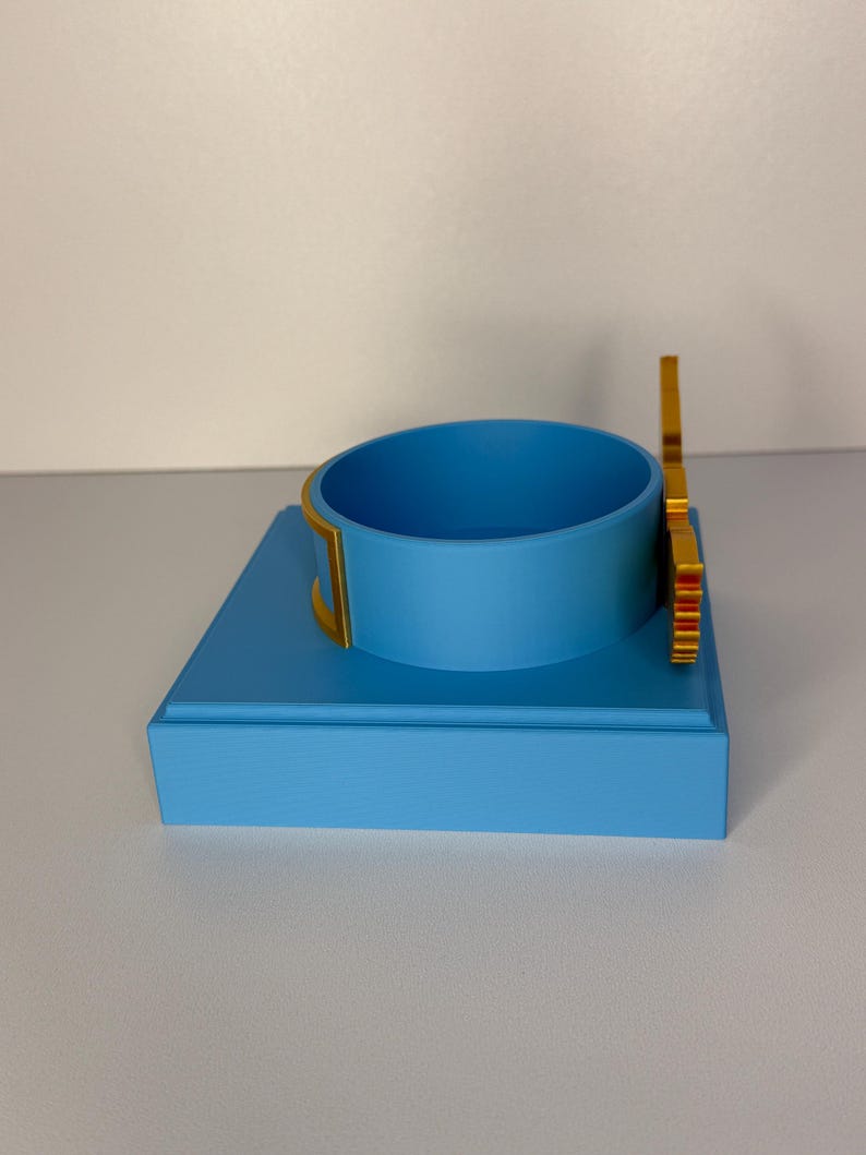 May include: A light blue square display base featuring a matching light blue circular ring holder in the center. The circular holder is accented with gold-colored trim on the sides and a vertical gold-colored decorative element attached to the back.