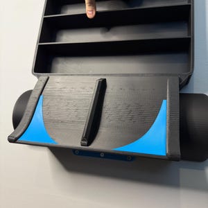 Op de afbeelding: Zwart, aan de muur gemonteerd displayrek met meerdere compartimenten. Het rek heeft een uniek ontwerp met blauwe accenten en een gebogen onderkant. De afbeelding toont het rek aan een lichtgrijze muur.