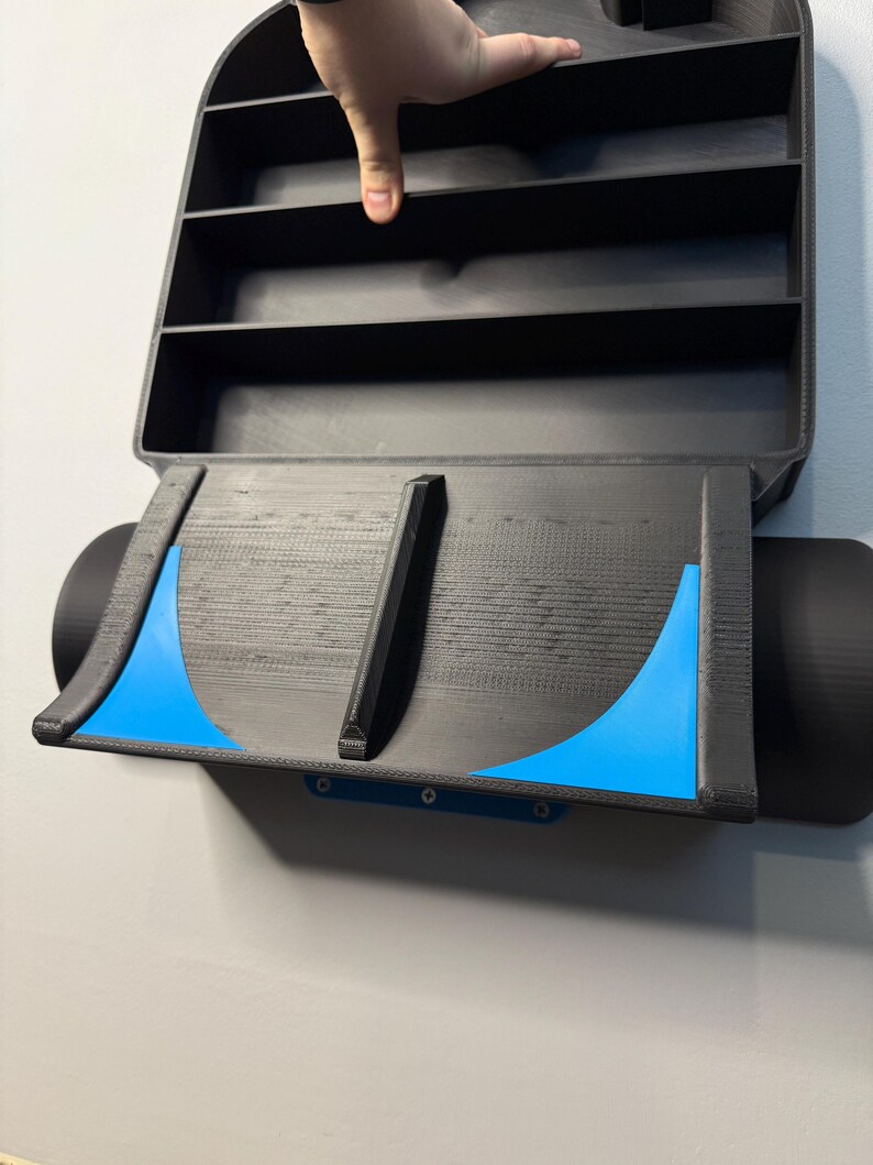 Op de afbeelding: Zwart, aan de muur gemonteerd displayrek met meerdere compartimenten. Het rek heeft een uniek ontwerp met blauwe accenten en een gebogen onderkant. De afbeelding toont het rek aan een lichtgrijze muur.