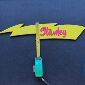 May include: A yellow arrow-shaped sign with the name "Stanley" in pink script. The sign is approximately 4 inches long. A green tape measure is used to measure the sign's length.