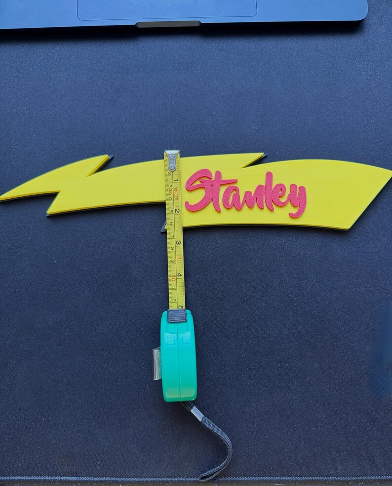 May include: A yellow arrow-shaped sign with the name "Stanley" in pink script. The sign is approximately 4 inches long. A green tape measure is used to measure the sign's length.