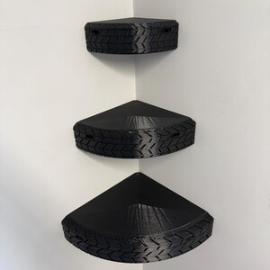 May include: Three black corner shelves mounted on a white wall. Each shelf is triangular with a textured tire tread design around the edges. The shelves are designed to fit snugly into a corner, maximizing space.
