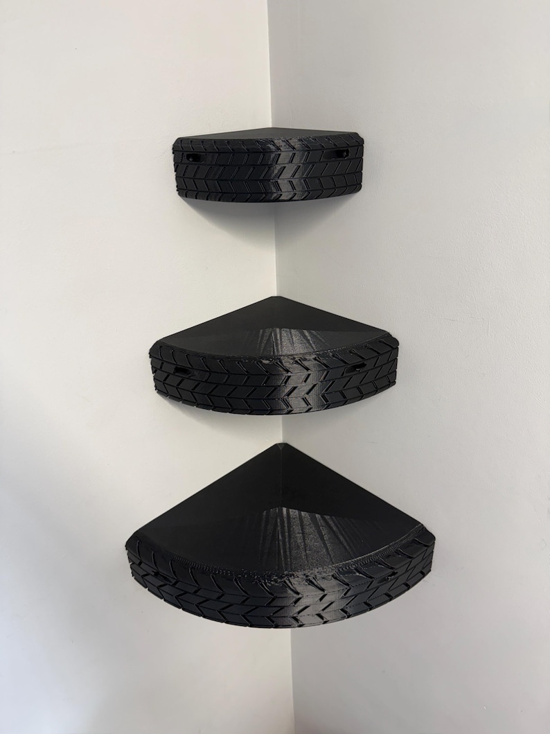 May include: Three black corner shelves mounted on a white wall. Each shelf is triangular with a textured tire tread design around the edges. The shelves are designed to fit snugly into a corner, maximizing space.
