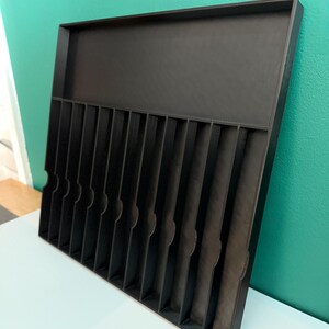 May include: A black organizer tray with multiple vertical dividers and a raised back section. The tray is designed to hold and separate items, with a textured surface and rounded cutouts on the dividers.