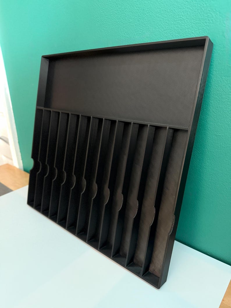 May include: A black organizer tray with multiple vertical dividers and a raised back section. The tray is designed to hold and separate items, with a textured surface and rounded cutouts on the dividers.