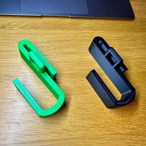 Two jumbo sized hooks for dog crates lay on a wooden desk. Green on the left and black on the right. They both have larger hook ends to allow for storing things such as long leads