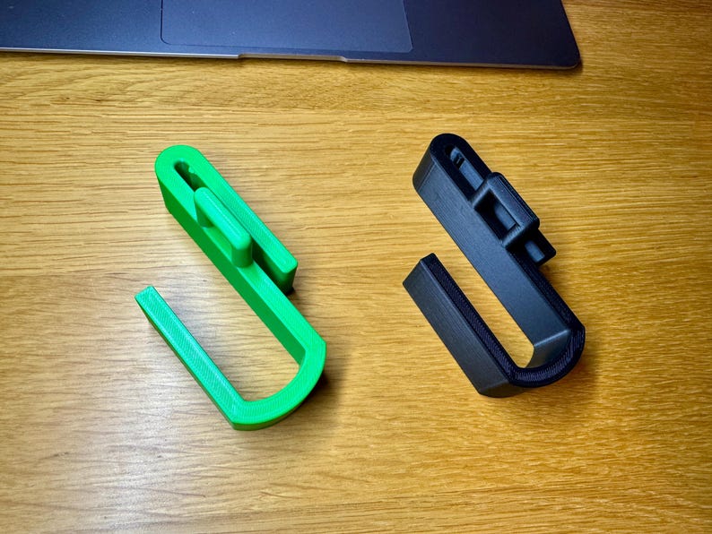Two jumbo sized hooks for dog crates lay on a wooden desk. Green on the left and black on the right. They both have larger hook ends to allow for storing things such as long leads