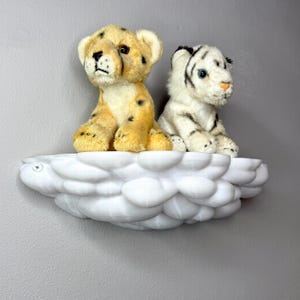 May include: A white cloud-shaped shelf holds two plush toy animals. One is a yellow cheetah cub with black spots, and the other is a white tiger cub with black stripes. The shelf is mounted on a gray wall.