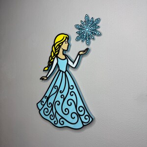 May include: A cartoon illustration of a woman in a light blue dress with a black outline, holding a blue snowflake. The woman has blonde hair in a braid. The dress has a decorative swirl design.