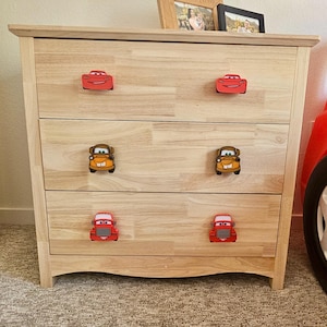 May include: A light wood dresser with three drawers, each adorned with car-shaped knobs in red, yellow, and orange. The dresser is set against a neutral wall and carpet.