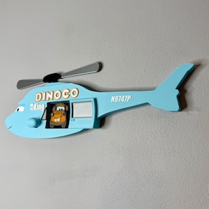 Puede incluir: Una decoraci&oacute;n de pared de madera en forma de helic&oacute;ptero azul claro con el texto DINOCO y King junto a un icono de corona. El helic&oacute;ptero tiene aspas plateadas, un peque&ntilde;o pomo redondo y una ventana recortada que muestra un cami&oacute;n marr&oacute;n.