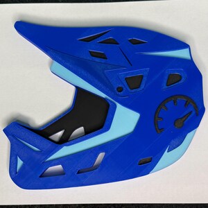 May include: A blue and light blue full-face helmet with a black interior. The helmet has a speedometer graphic on the side and several ventilation openings. The helmet is designed for sports like mountain biking or downhill racing.
