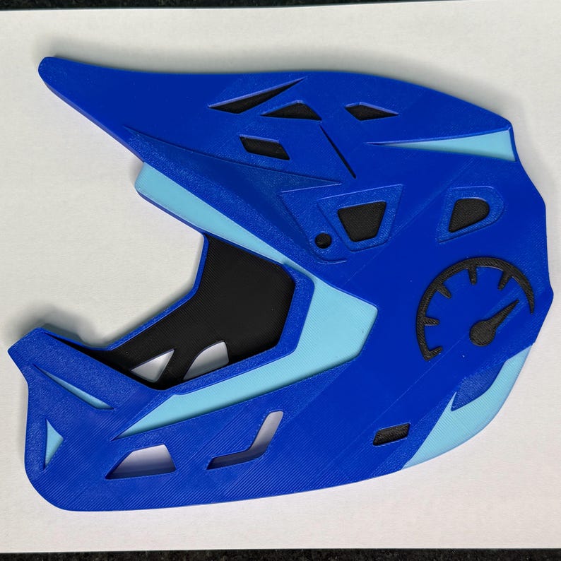May include: A blue and light blue full-face helmet with a black interior. The helmet has a speedometer graphic on the side and several ventilation openings. The helmet is designed for sports like mountain biking or downhill racing.