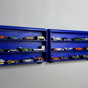 Two blue wall-mounted toy car display shelves connected to a customisable 3D-printed train engine labelled “Noah.” The shelves are filled with die-cast vehicles and designed to look like train carriages.