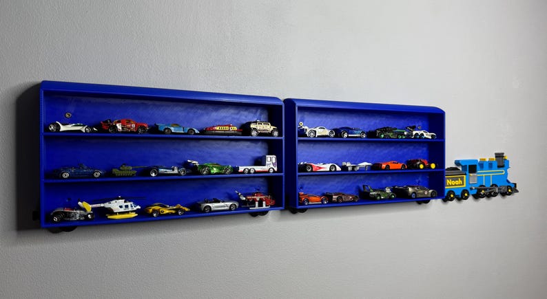 Two blue wall-mounted toy car display shelves connected to a customisable 3D-printed train engine labelled “Noah.” The shelves are filled with die-cast vehicles and designed to look like train carriages.