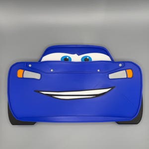 May include: A blue car-shaped pillow with a smiling face, headlights, and black tires. The pillow has white eyes with blue irises and a white mouth. The car's body is a vibrant blue color.