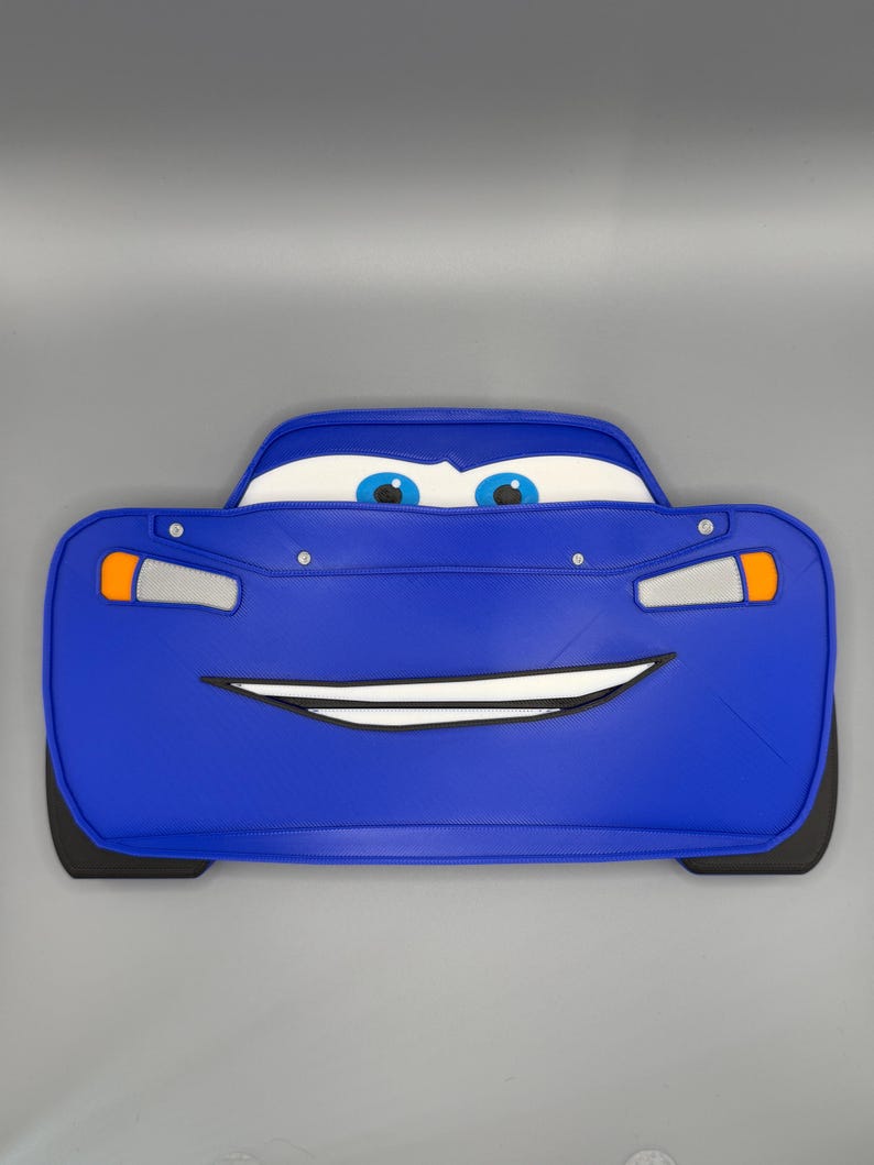 May include: A blue car-shaped pillow with a smiling face, headlights, and black tires. The pillow has white eyes with blue irises and a white mouth. The car's body is a vibrant blue color.