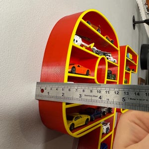 May include: A red and yellow shelf in the shape of the word "HOT" is filled with miniature toy cars. The shelf is approximately 5 inches wide. The cars are various colors and styles.