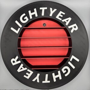 May include: A circular black object with the word "LIGHTYEAR" in white lettering. The center features a red, slatted design. A small, dark screw is visible at the top. The overall design is reminiscent of a spaceship element.