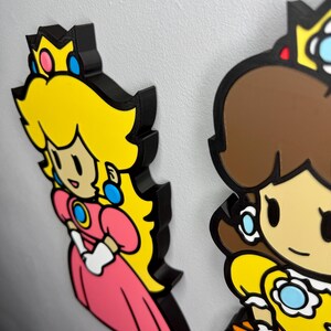 Op de afbeelding: Twee kleurrijke karakteruitsparingen, prinses Peach en prinses Daisy, uit de Super Mario-videogameserie. Peach draagt een roze jurk en Daisy een gele jurk. Beide hebben zwarte contouren en zijn op een wit oppervlak gemonteerd.