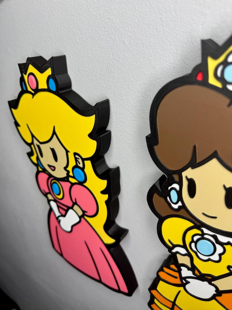 Op de afbeelding: Twee kleurrijke karakteruitsparingen, prinses Peach en prinses Daisy, uit de Super Mario-videogameserie. Peach draagt een roze jurk en Daisy een gele jurk. Beide hebben zwarte contouren en zijn op een wit oppervlak gemonteerd.