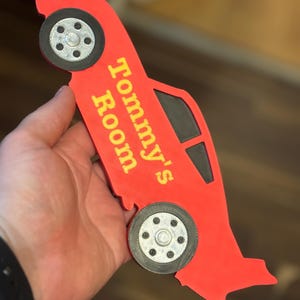 May include: A red car-shaped sign with the text "Tommy's Room" in yellow. The sign has black wheels and windows. The sign is held in a hand.