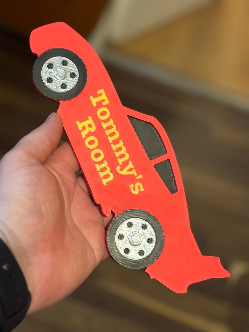 May include: A red car-shaped sign with the text "Tommy's Room" in yellow. The sign has black wheels and windows. The sign is held in a hand.