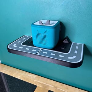 May include: A teal speaker with a perforated design is on a road-themed shelf. The shelf is black and grey with white road markings and a chequered flag. The speaker has grey ear-shaped accents on top.